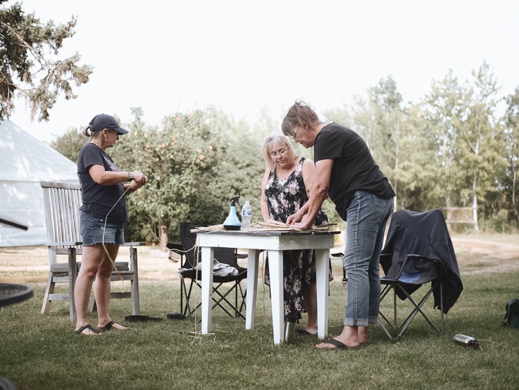 Three weavers working together around a small white table on the grass, one measuring out cord, with a tent and poplars behind them.