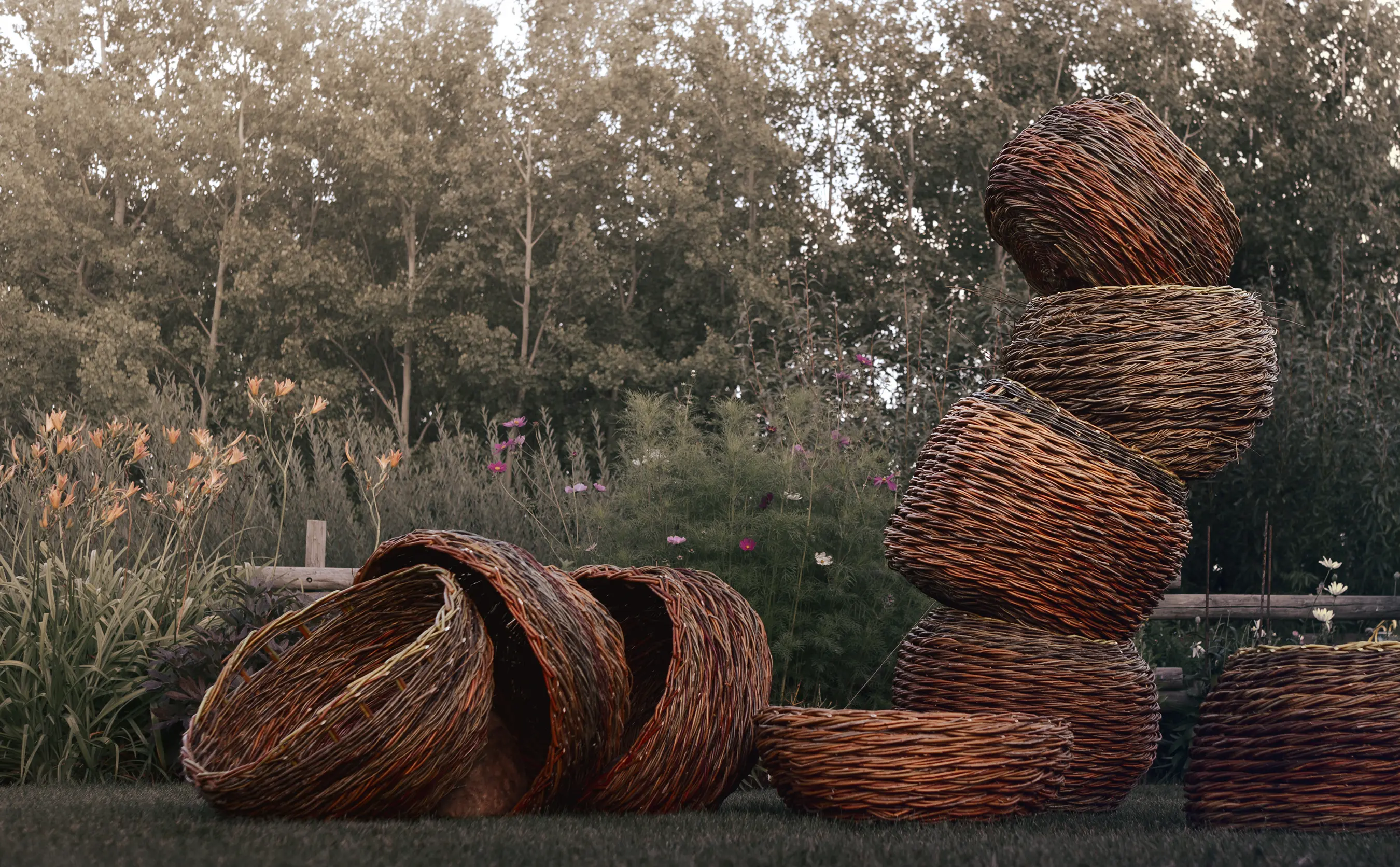 Large sculptural willow baskets standing and lying on the grass in front of a garden bed of orange lilies and cosmos.